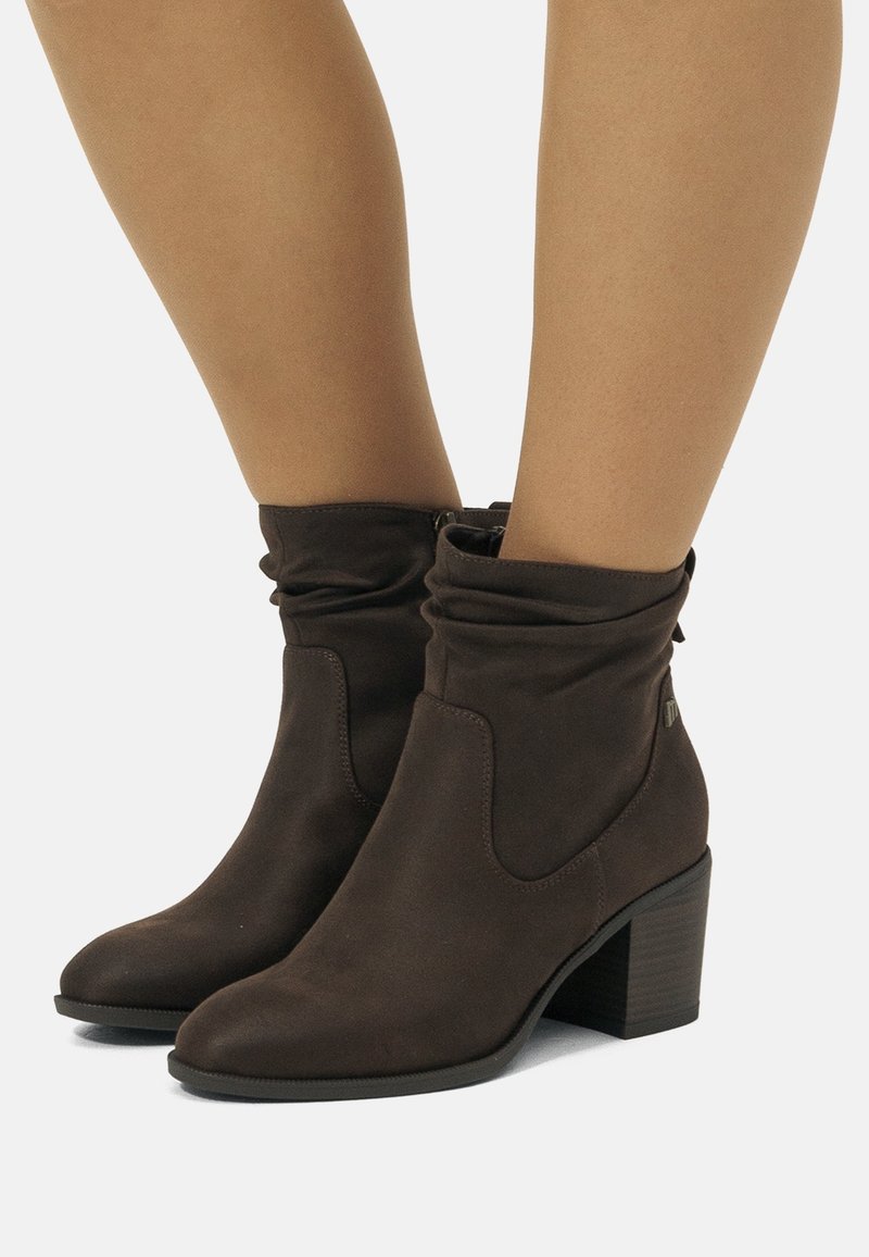 Bottines en daim marron à tige plissée et talons carrés, portées sur jambes nues sur un fond clair uni.