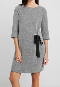 Robe texturée grise à manches trois-quarts, poche latérale, et détail de lien ajustable noir à la taille. Encolure ronde simple.