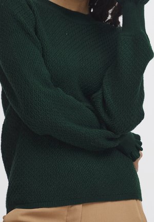 Žena v tmavě zeleném texturovaném pleteném svetru s dlouhými rukávy a béžových kalhotách na světlém pozadí.