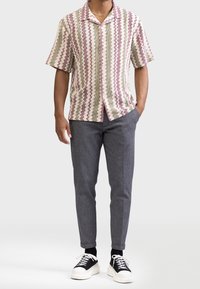 Chemise à manches courtes avec un motif en zigzag crème, bordeaux et vert, associée à un pantalon gris ajusté et des baskets noires à semelles blanches.