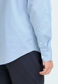 Cămașă albă cu un model albastru deschis și un mic design floral, cu manșete cu nasturi și un tiv curbat. Purtați-o cu pantaloni închiși la culoare, arătând o parte din mânecă.