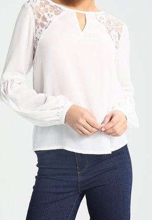Femme portant un chemisier blanc à manches longues avec un empiècement en dentelle et un col en trou de serrure, associé à un jean bleu foncé moulant, mains jointes à la taille.