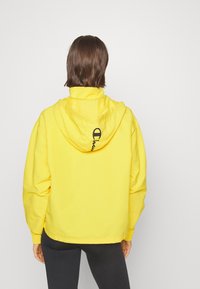 Gul huva jacka gjord av lättviktsmaterial med elastisk nederkant, med en svart logotyp på ryggen och en justerbar huva.