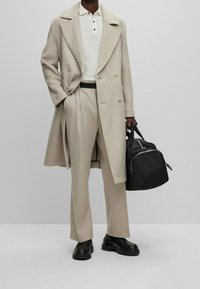 Manteau en laine beige sur polo blanc, pantalons beige à jambes larges et chaussures noires, tenant un petit sac noir avec des accents métalliques argentés.