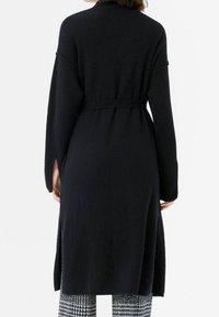 Lång svart kofta i mjukt material, med midjebälte, vida ärmar och sidofack för bättre rörlighet.