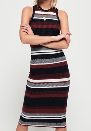 Femme portant une robe sans manches, longueur genou, avec des rayures horizontales noires, blanches, grises et rouge foncé, les bras croisés, portant deux colliers en or.