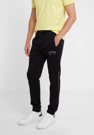 Mand iført sorte Hugo Boss joggingbukser med gyldt logo og hvide sneakers, stående med den ene hånd i lommen.