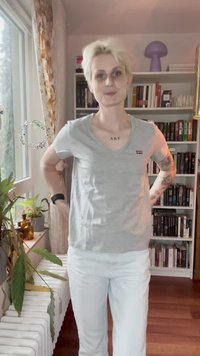 Person with short blonde hair, grey t-shirt, white pants, and tattooed arms standing indoors by a window and bookshelf with plants nearby.