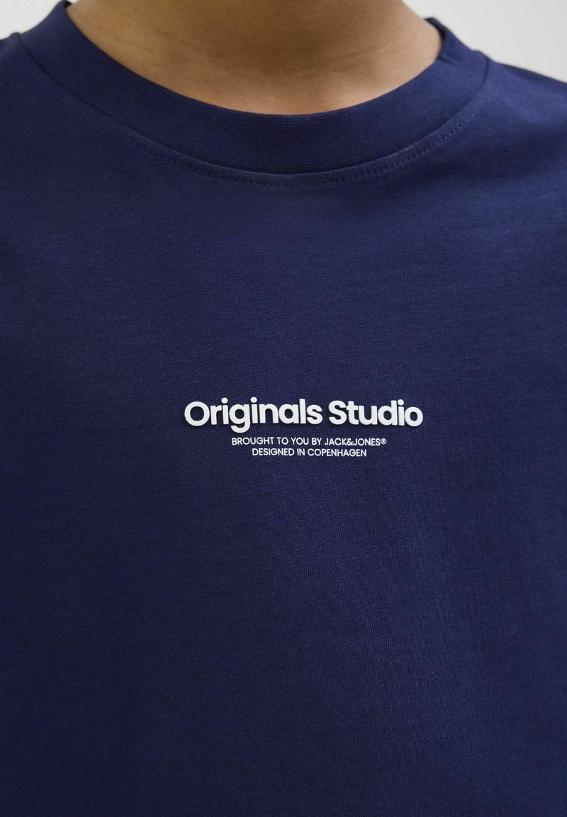 Námorně modré bavlněné tričko s kulatým výstřihem; obsahuje bíle potisknutý text "Originals Studio" s dalším brandingem níže.