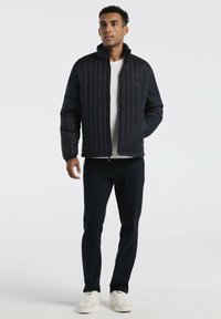 Man in een zwart gewatteerd jack over een wit shirt, zwarte broek en witte sneakers, staand met één hand in de zak van het jack.