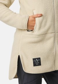 Giacca in pile di colore crema, con tasche laterali, chiusura con zip e una patch nera con logo all'orlo. Materiale morbido e strutturato.