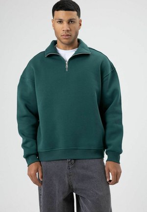 Dunkelgrüner Pullover mit halbhohem Reißverschluss, lockerer Passform, gerippten Bündchen und Saum, aus weichem, dickem Material. Getragen über grauen Jeans.