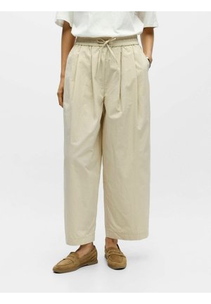 Oseba, oblečena v ohlapne bež priložnostne hlače z žepi in rjavimi loaferji, stoji pred enobarvnim belim ozadjem.