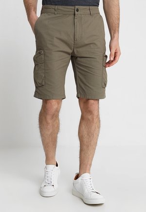 Homme portant un short cargo vert olive avec poches latérales et des baskets blanches, debout sur un sol blanc contre un fond uni.