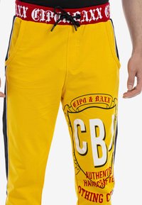 Gelbe Jogginghose mit einem roten elastischen Bund und schwarzem Kordelzug sowie seitlichen Streifen. Großes bedrucktes grafisches Design in mehreren Farben.