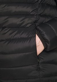 Doudoune noire matelassée avec motif en losanges, dotée d'une poche latérale et d'une texture lisse en nylon. Le détail de la fermeture éclair est visible.