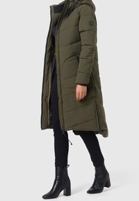 Person i en olivgrön lång vadderad jacka med dragkedjor, svarta byxor och svarta ankle boots med klack framför en neutral bakgrund.