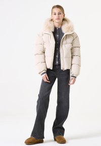 Beige Daunenjacke mit Kunstfellkragen, Reißverschluss vorne, gestepptem Design, getragen über einem grauen Button-up-Pullover und dunklen Jeans. Beigefarbene Hausschuhe runden das Outfit ab.