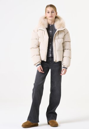 Chaqueta acolchada beige con cuello de pelo sintético, cierre con cremallera, diseño acolchado, llevada sobre un suéter gris de botones y jeans oscuros. Zapatillas color crema completan el atuendo.