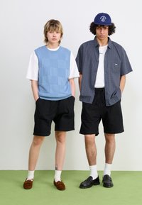 Zwei Personen stehen nebeneinander. Der eine trägt ein hellblaues strukturiertes Weste über einem weißen T-Shirt, schwarze Shorts und braune Schuhe. Der andere trägt ein kariertes Hemd, schwarze Shorts und schwarze Schuhe.