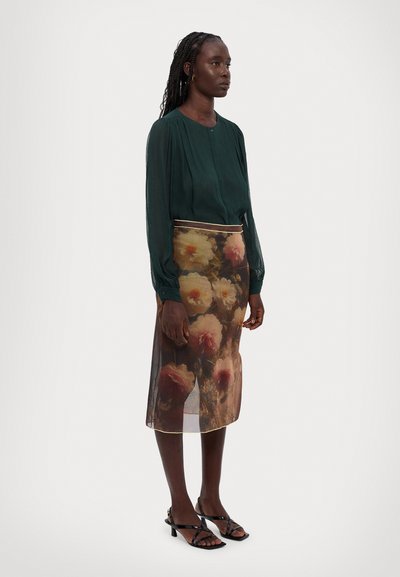 Grøn, langærmet bluse med plisseringer parret med en gennemsigtig, blomstertrykt nederdel med store, lyse blomster og en kontrasterende kant.