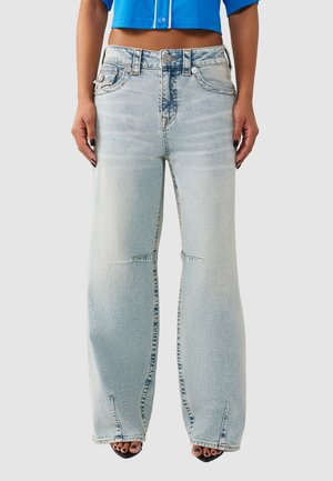 Femme portant un jean large bleu clair et un haut court bleu à boutons, debout avec les bras détendus le long du corps.