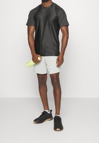 Camiseta deportiva negra con paneles laterales de malla, combinada con pantalones cortos gris claro y zapatillas negras. El modelo sostiene una botella de agua verde.