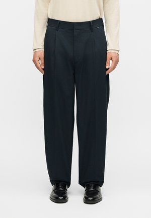 Άνδρας που φοράει μαύρο παντελόνι με φαρδιά μπάσκα και μαύρα επίσημα παπούτσια, στέκεται μπροστά σε ένα απλό λευκό φόντο.