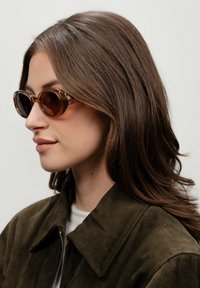 Gafas de sol redondas marrones con un marco transparente, que presentan un acabado brillante. Llevadas con una chaqueta de gamuza verde oscuro, mostradas de perfil lateral.