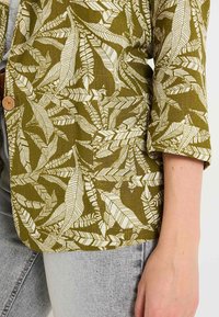 Blazer vert à motif fabriqué en tissu texturé, présentant un design de feuilles blanches. Détail de bouton unique à l'avant et coupe décontractée.