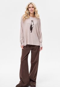 Jasnoróżowa koszula z długim rękawem z nadrukiem, zestawiona z luźnymi brązowymi spodniami o swobodnym kroju i fakturowanej tkaninie.