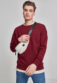 Burgunderfarget genser med ribbestrikket rund hals og mansjetter, kombinert med en lysfarget undertrøye. En hvit teksturert crossover-veske med svart stropp.