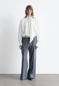 Weiße Bluse mit hochgeschlossenem Kragen und Schleife, kombiniert mit weit geschnittenen grauen Hosen. Glattes Material, lockerer Sitz und lange Ärmel.