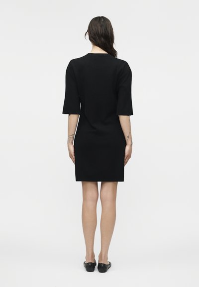 Γυναίκα με σκούρα μαλλιά, φορώντας μαύρο φόρεμα μήκους μέχρι το γόνατο με μανίκια μέχρι τον αγκώνα και μαύρα επίπεδα παπούτσια, στέκεται γυρισμένη προς τα πίσω σε απλό φόντο.