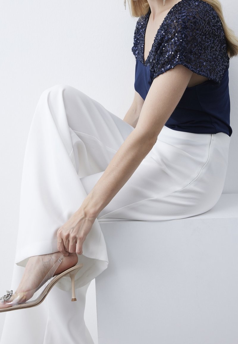 Femme assise, une jambe croisée, portant un pantalon blanc large, un haut bleu marine à sequins et des chaussures à talons hauts à brides transparentes avec décoration.