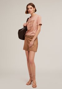 Vaaleanpunainen t-paita, jossa on teksturoitu graafinen kuvio, yhdistettynä vaaleanruskeisiin shortseihin ja nudenvärisiin sandaaleihin, joissa on hihnat. Käsissä musta laukku.