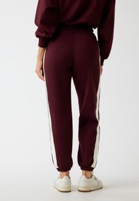 Bordeaux joggings met witte zijstreep, elastische tailleband en boorden, gemaakt van een zachte stof. Draag ze met witte sneakers.
