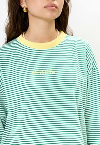 Groen en witte gestreepte longsleeve top met een gele ribbelhalslijn, voorzien van een gele logotekst in het midden van de borst.