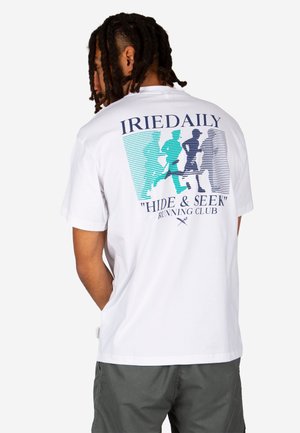 Mann mit Dreadlocks, der ein weißes T-Shirt mit blauen und türkisfarbenen Laufmotiven und dem Text „IRIEDAILY HIDE & SEEK RUNNING CLUB“ auf dem Rücken trägt.