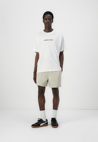 Camiseta blanca de algodón con texto rojo, pantalones cortos beige y zapatillas negras con acentos grises. El modelo está de pie contra un fondo liso.