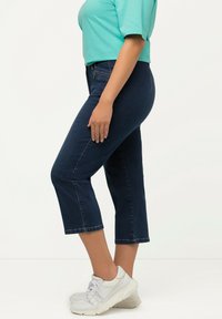 Tummansiniset lyhyet farkut suoralinjaisella leikkauksella, valmistettu denimistä, yhdistettynä teal-väriseen paitaan ja valkoisiin lenkkareihin. Tekstuuri näyttää sileältä.