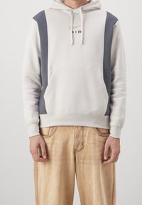 Hoodie med en ljus creamfärgad kropp, mörkgrå sidopaneler, framficka i kängurustil och en guldsymbol. Bärs med beiga byxor.