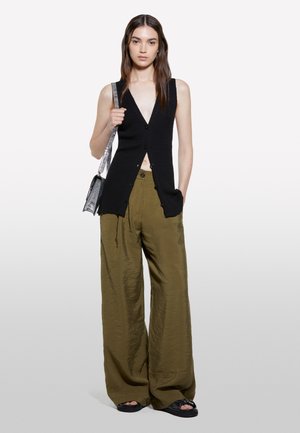 Giovane donna che indossa un gilet nero senza maniche con bottoni, pantaloni larghi verde oliva, sandali neri e porta una borsa a tracolla argentata.