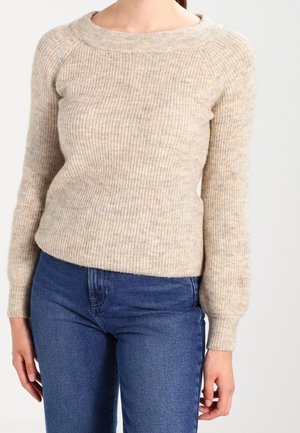 Vrouw draagt een beige geribbelde gebreide trui met lange mouwen en blauwe spijkerbroek tegen een effen achtergrond.