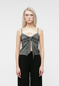 Νεαρή γυναίκα με μακριά ξανθά μαλλιά, φοράει ένα γκρίζο αμάνικο τοπ με κόμπο μπροστά και μαύρο φαρδύ παντελόνι, στέκεται μπροστά σε ένα απλό φόντο.