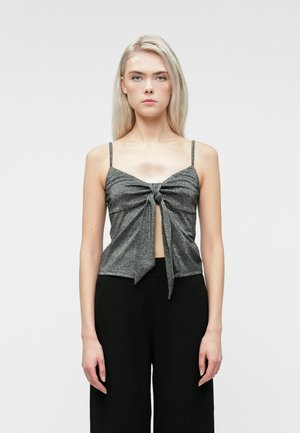 Młoda kobieta z długimi blond włosami nosi szary bezrękawnik z węzłem z przodu i czarne szerokie spodnie, stojąc na jednolitym tle.