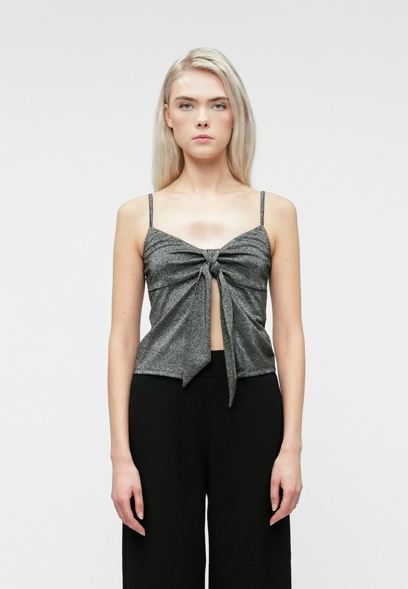 Νεαρή γυναίκα με μακριά ξανθά μαλλιά, φοράει ένα γκρίζο αμάνικο τοπ με κόμπο μπροστά και μαύρο φαρδύ παντελόνι, στέκεται μπροστά σε ένα απλό φόντο.