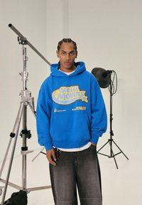 Blå sweatshirt med en stor gul grafisk text "Still Moving." Har en huva, ribbade ärmslut och en avslappnad passform. Modellen har mörka jeans.