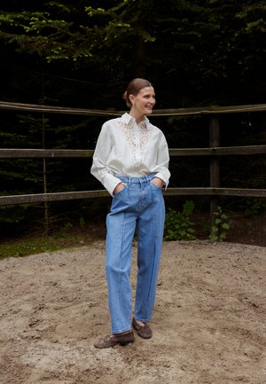 Kvinde smilende udendørs iført hvid blonderbluse, højtaljede blå jeans og brune loafers, stående på sandet grund ved et træhegn.