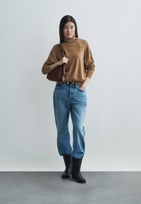 Pruun kudumistest kõrge kaelusega kampsun, lahtine istuvus; kõrge vöökohaga sinised teksad; mustad põlveni ulatuvad saapad; pruuni õlakotiga käekott.